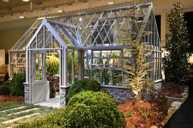 Glass greenhouse with white trim and stone base, featuring an open door and lush greenery inside and out.