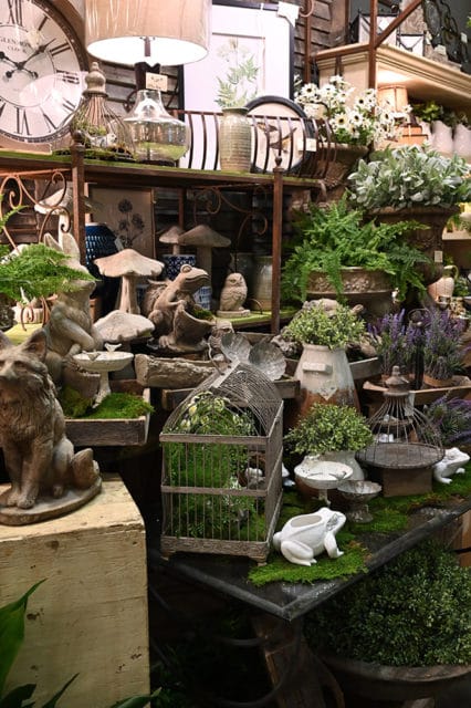 Display of garden-themed decor including a large clock, ceramic frogs, mushroom statues, birdcages, and potted plants.
