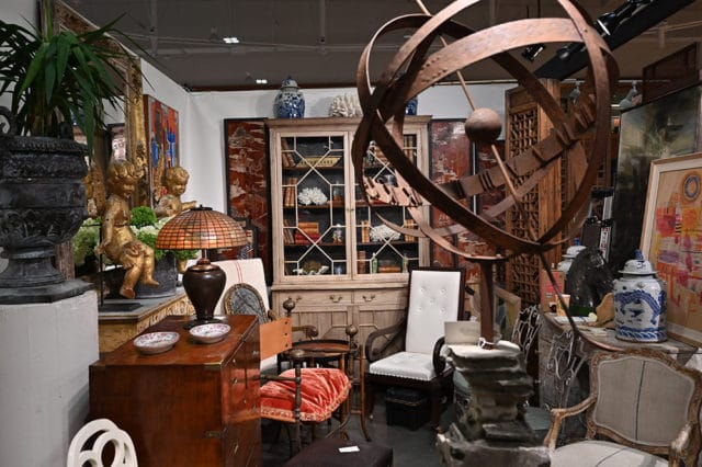 Antique shop interior with a large metal armillary sphere, a glass-fronted cabinet filled with books, and various decorative items including statues and porcelain vases.