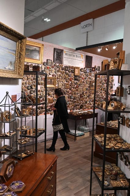 Woman browsing a display of hexagonal and square decorative items, many with gold accents, on a wall and shelves. A sign reads 'MUSEUM BEES'.