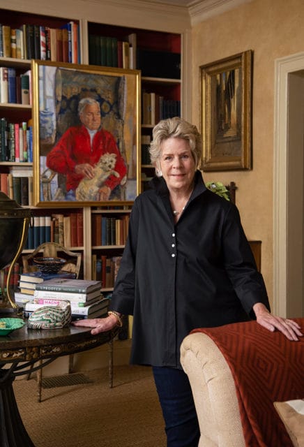 Woman in a black collared shirt and jeans stands in a room with bookshelves, a painting of a man and dog, and a draped throw on a chair.