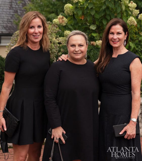 Three women in black dresses stand outside with greenery behind them.