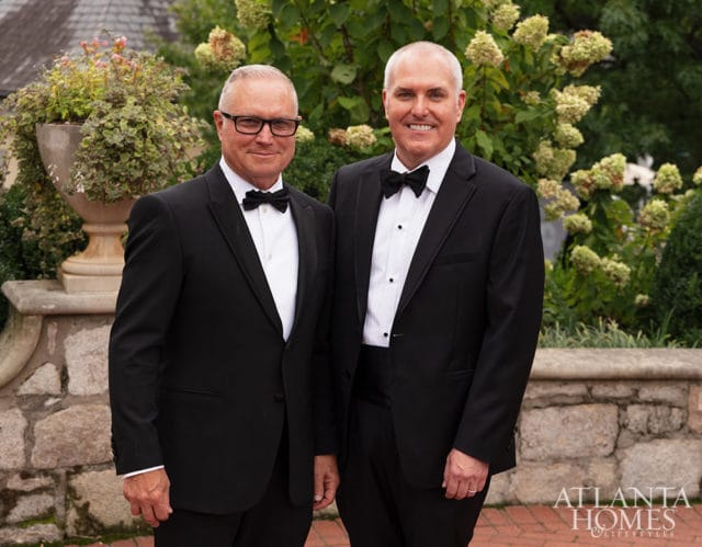 Two men in tuxedos standing outdoors with hydrangeas and stone wall behind them.