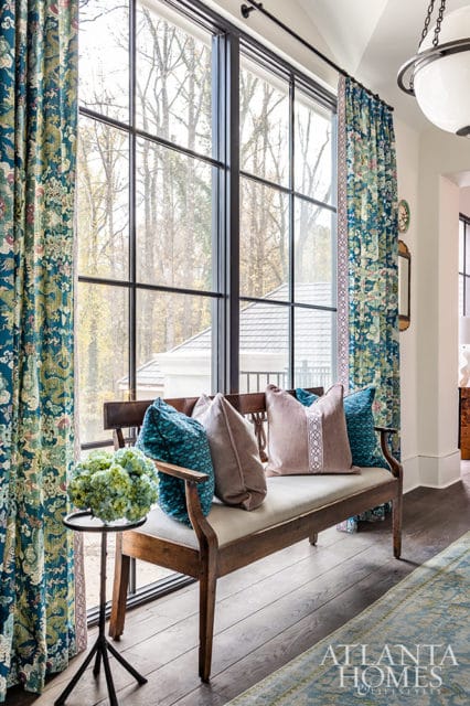 Wooden bench with teal and taupe pillows in front of large window with patterned curtains.