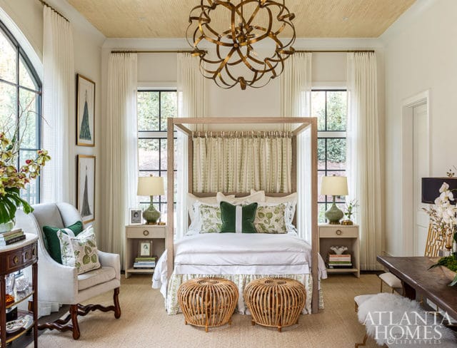 Bedroom with canopy bed, green floral pillows, rattan stools, and grasscloth ceiling.