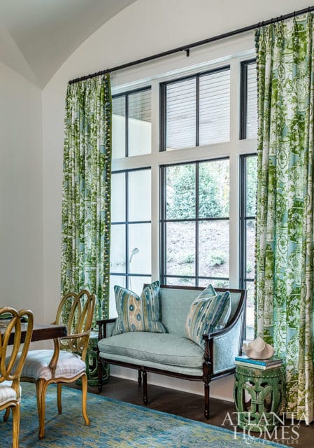 Light blue settee with patterned pillows next to a large window with green floral curtains.