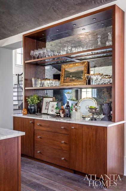Wooden bar cabinet with mirrored backsplash displaying glassware, framed art, and barware. Marble countertop and dark wood flooring visible.