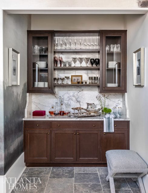 Dark wood bar cabinet with marble countertop and glass-front upper cabinets filled with glassware.