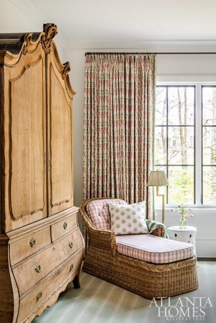 Wicker chaise with pink gingham cushions and floral pillows next to antique armoire.