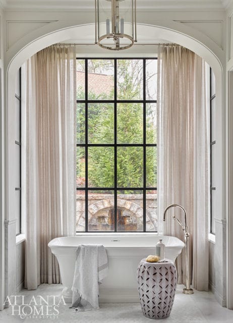 Freestanding white bathtub with a towel, next to a decorative stool and a tall faucet, in front of