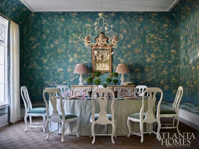 Dining room with teal floral wallpaper, white chairs with green seats, and a patterned tablecloth.