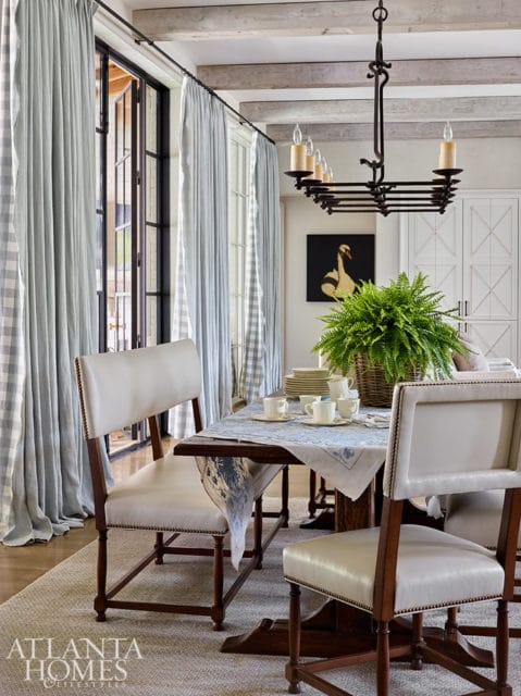 Dining room with light blue gingham curtains, a dark wood table set with white dishes and a fern centerpiece, and upholstered chairs.