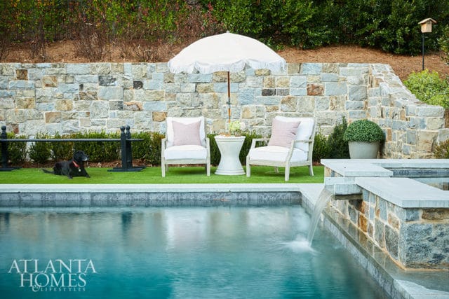 Stone wall with two white chairs, umbrella, and black dog by pool