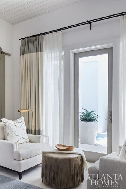 Living room with white armchair, fringed ottoman, and floor-to-ceiling curtains.