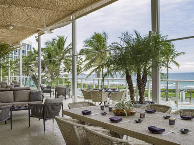 Dining table with place settings and wicker chairs, overlooking palm trees and the ocean.
