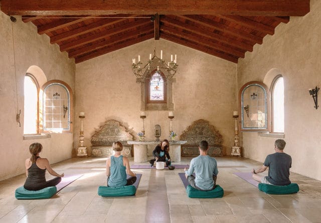 People meditating in a room with arched windows, exposed wooden beams, and ornate wall decor.