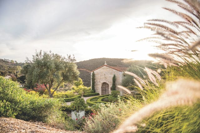 Stone chapel with arched wooden doors, surrounded by manicured gardens and olive trees.