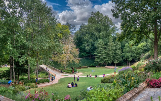 People relaxing in a park with lush green grass, trees, and colorful flowers under a cloudy sky.