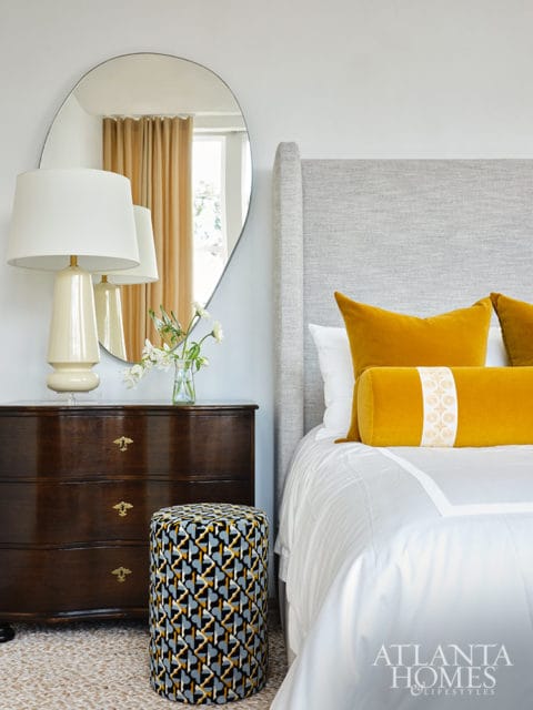 Bedroom with gray upholstered headboard, yellow pillows, dark wood dresser, and patterned ottoman.