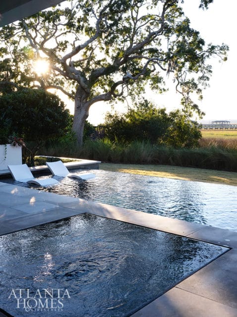 Infinity pool with submerged lounge chairs and spa overlooking marshland and large oak tree