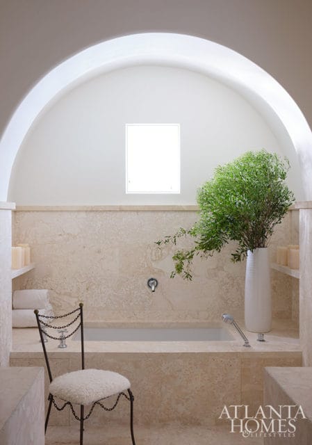 Travertine tiled bathroom with arched ceiling, tub, white vase with greenery, and metal chair.