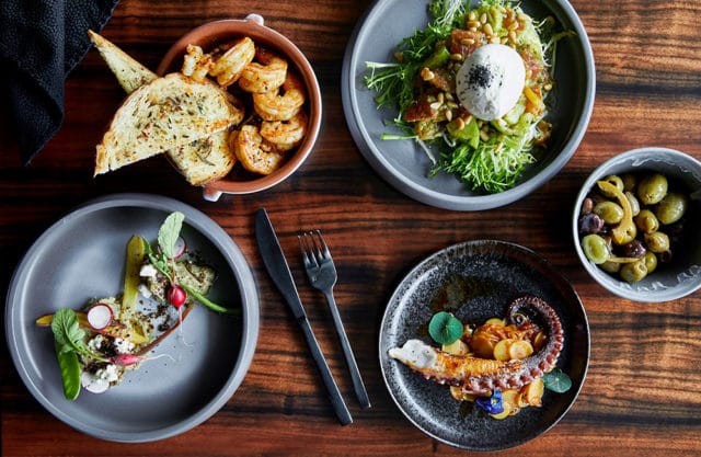 Four plates of food and a bowl of olives on a wooden table.