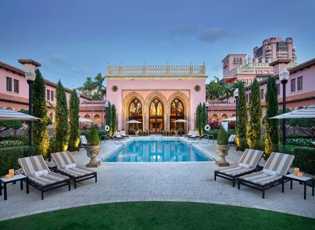 Pink hotel with arched windows and a swimming pool, surrounded by cypress trees and lounge chairs.