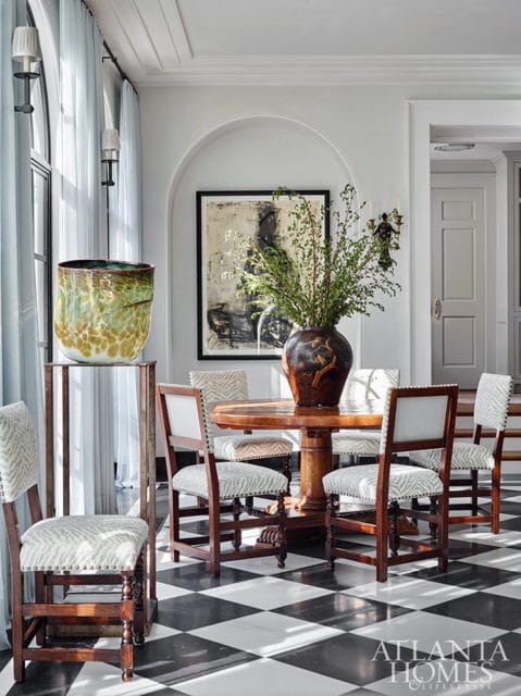 Dining room with round wooden table, upholstered chairs, abstract art, and large glass vase.