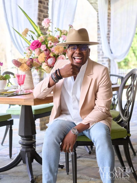 Man in tan hat and peach blazer sitting at a table with floral arrangement.