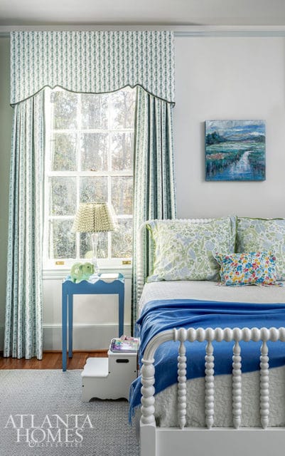 Bedroom with white spindle bed, blue blanket, floral pillows, and patterned curtains.