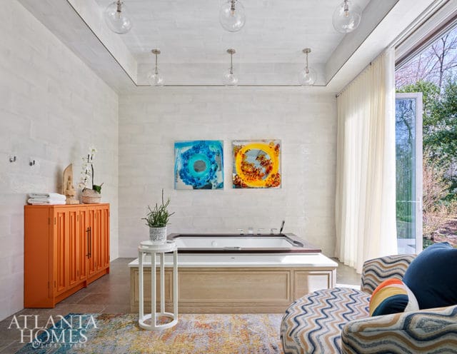 Modern bathroom with white brick walls, built-in tub, orange cabinet, and abstract art.