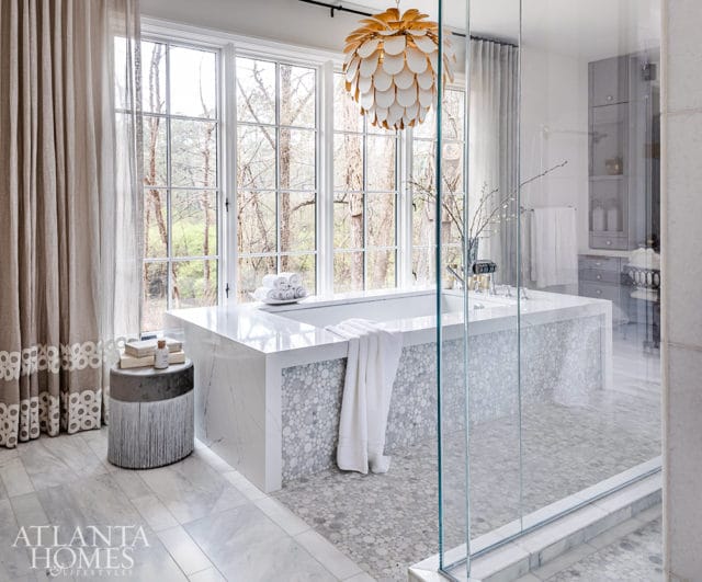 White marble bathtub with pebble tile surround, large windows, and gold chandelier.