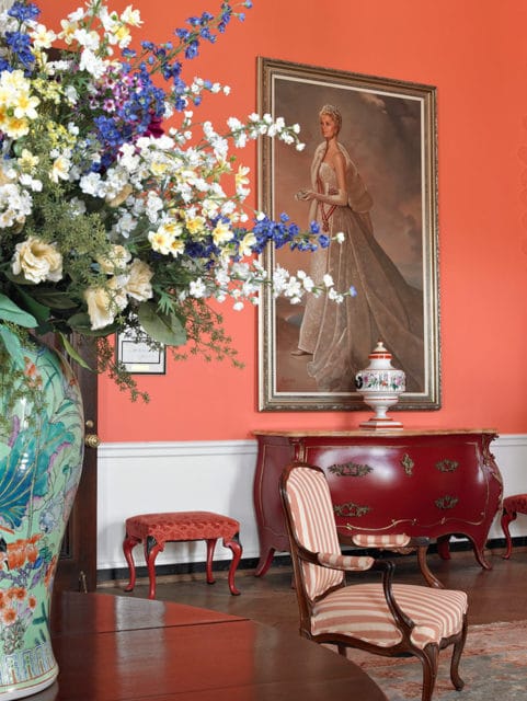 Coral walls with a portrait, red dresser, striped armchair, and floral arrangement