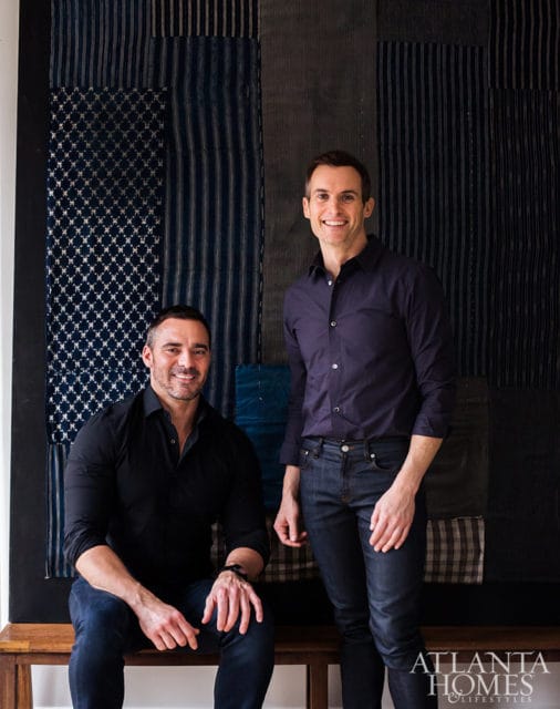 Two men smiling in front of a textile wall hanging.