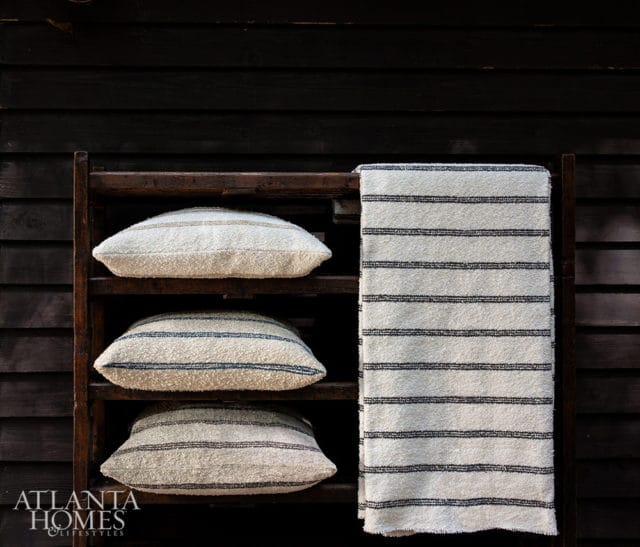 Three textured pillows and a striped towel on a wooden shelf against a dark wood wall.