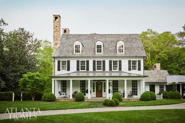 White farmhouse with stone chimney, wraparound porch, and manicured lawn.