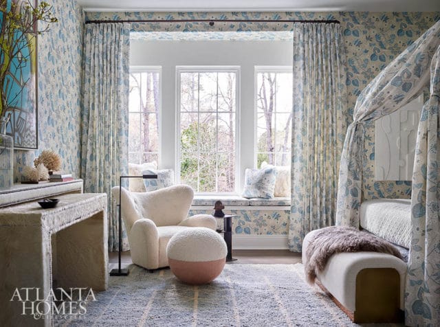 Bedroom with blue floral wallpaper, white armchair, ottoman, and canopy bed.