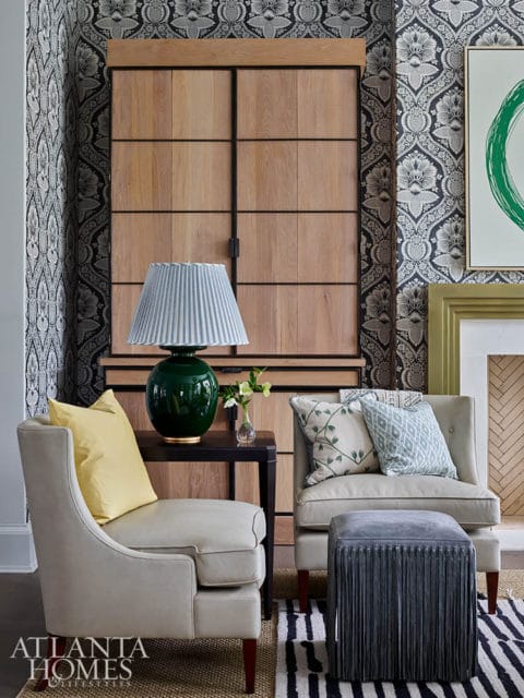 Living room with patterned wallpaper, wood cabinet, two chairs, and fringed ottoman.
