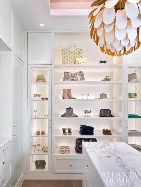 White walk-in closet with shelves displaying hats, shoes, bags, and sunglasses. Marble island in