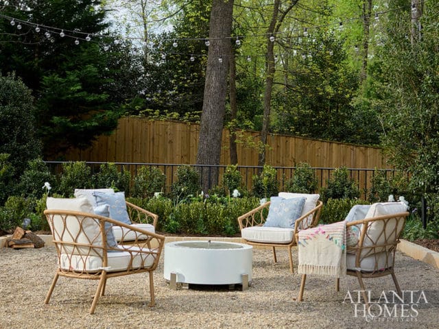 Outdoor seating area with rattan chairs and a white fire pit on a gravel patio, surrounded by lush