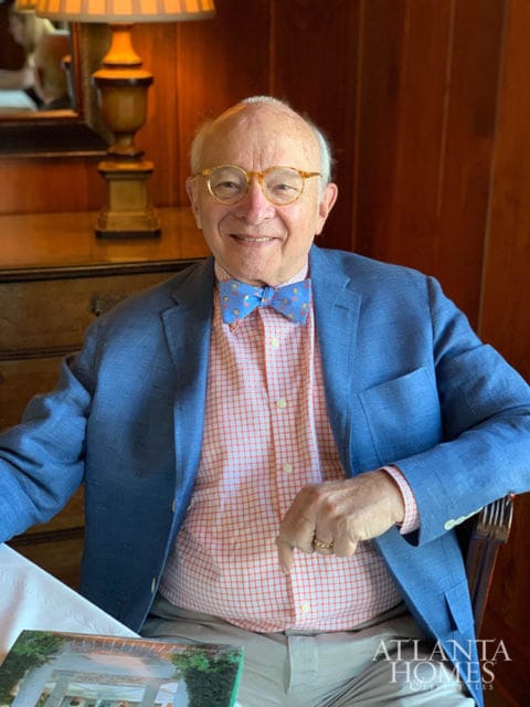 Elderly man with glasses and a bow tie smiles, wearing a blue blazer over an orange checkered shirt.
