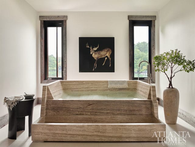 Travertine bathtub with tiered design, artwork of deer above, and a potted tree beside it.
