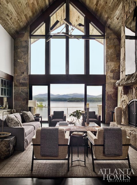 Living room with stone fireplace, wood-paneled ceiling, and large windows overlooking a lake.