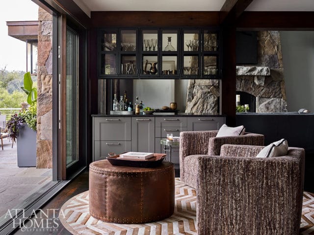 Living room with gray cabinets, glass-fronted bar, stone fireplace, and upholstered chairs.