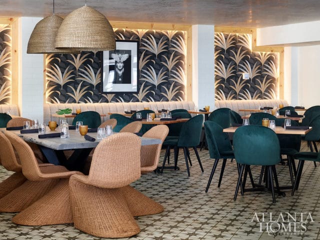Restaurant dining room with palm leaf wallpaper, wicker chairs, and green velvet chairs.
