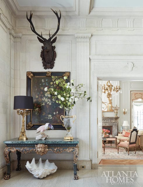 Hallway with mounted deer head, floral painting, ornate table, and doorway to living room.