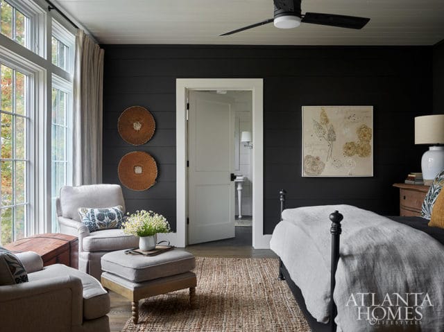 Bedroom with dark shiplap walls, light gray armchair and ottoman, and a bed with light gray linens.