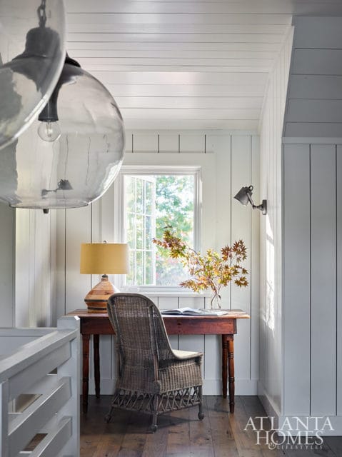 Small desk with wicker chair, lamp, and vase of fall leaves. Shiplap walls and wood floor.