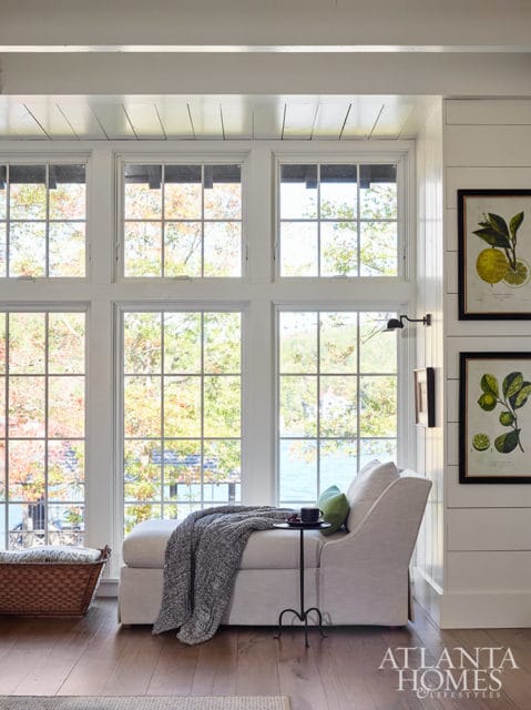 Light-colored chaise lounge with gray throw blanket and green pillow, by large windows overlooking