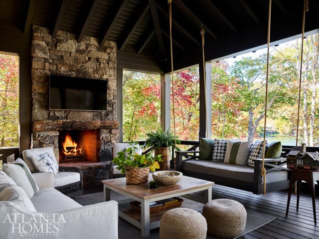 Covered porch with stone fireplace, seating, and swing overlooking a lake in fall.